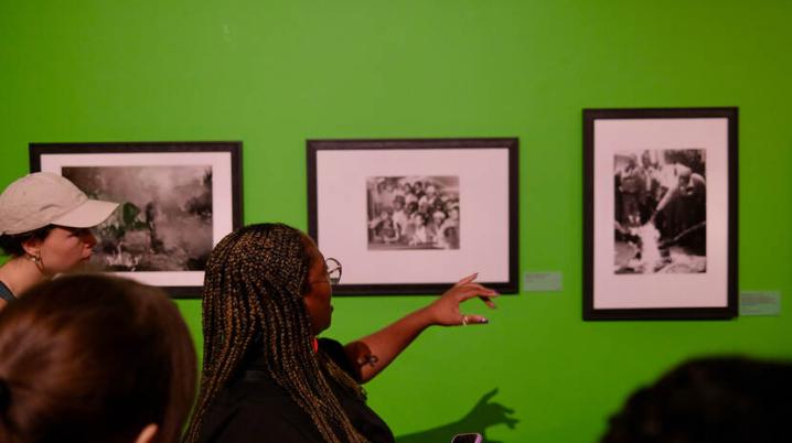 woman wearing big round glasses points at black and white photographs hung in black frames against a bright green wall