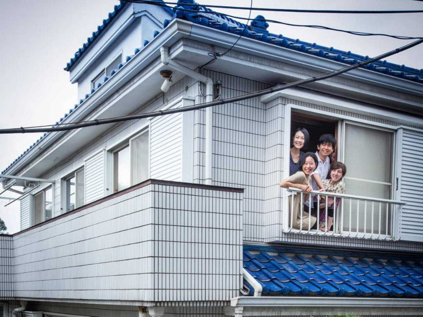 Four people leaning out of the window of a white house with a blue roof at Co.iki residency in Tokyo, Japan