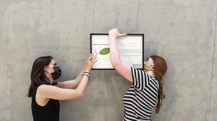 two women wearing facemasks holding an art work against a grey wall