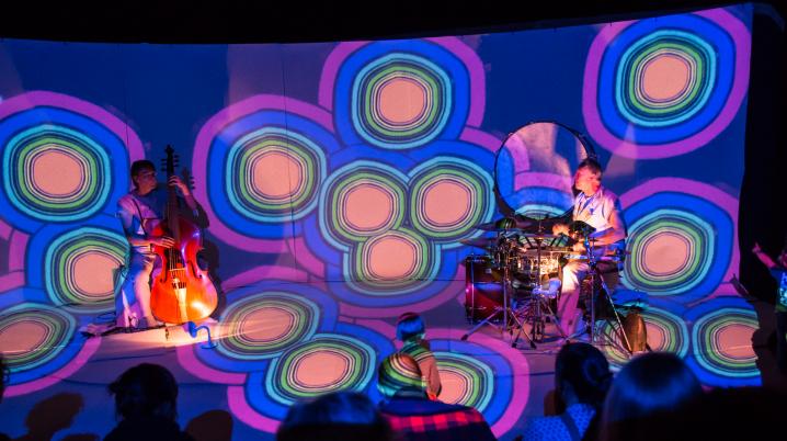 a colourful stage with a cello player and a drummer and a young audience in the foreground
