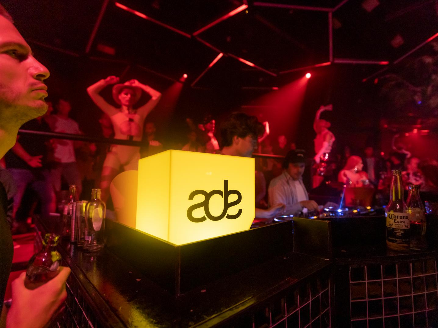 ADE logo on a DJ deck surrounded by people dancing