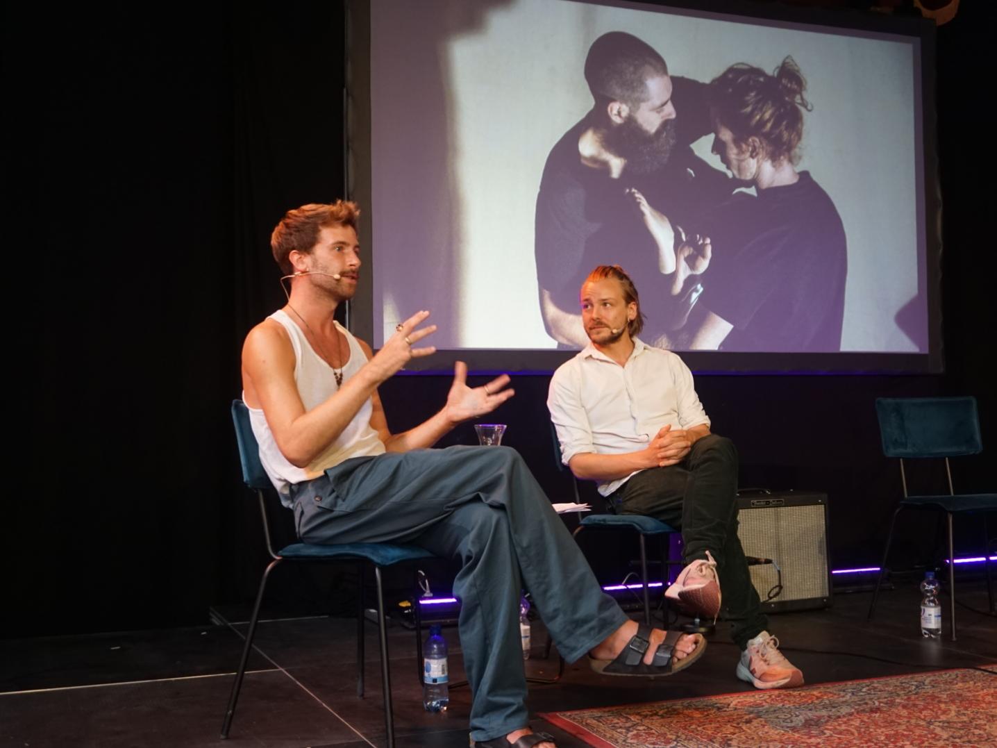 Two people, Philipp Cahrpit and Simon de Leeuw, on stage, with a still from Cahrpit's performance on the background.