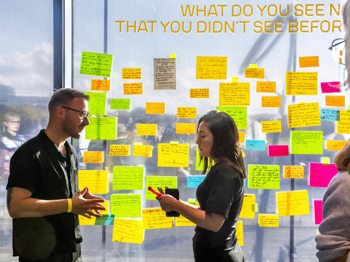 two people standing in front of a glass wall with lots of coloured sticky notes on it