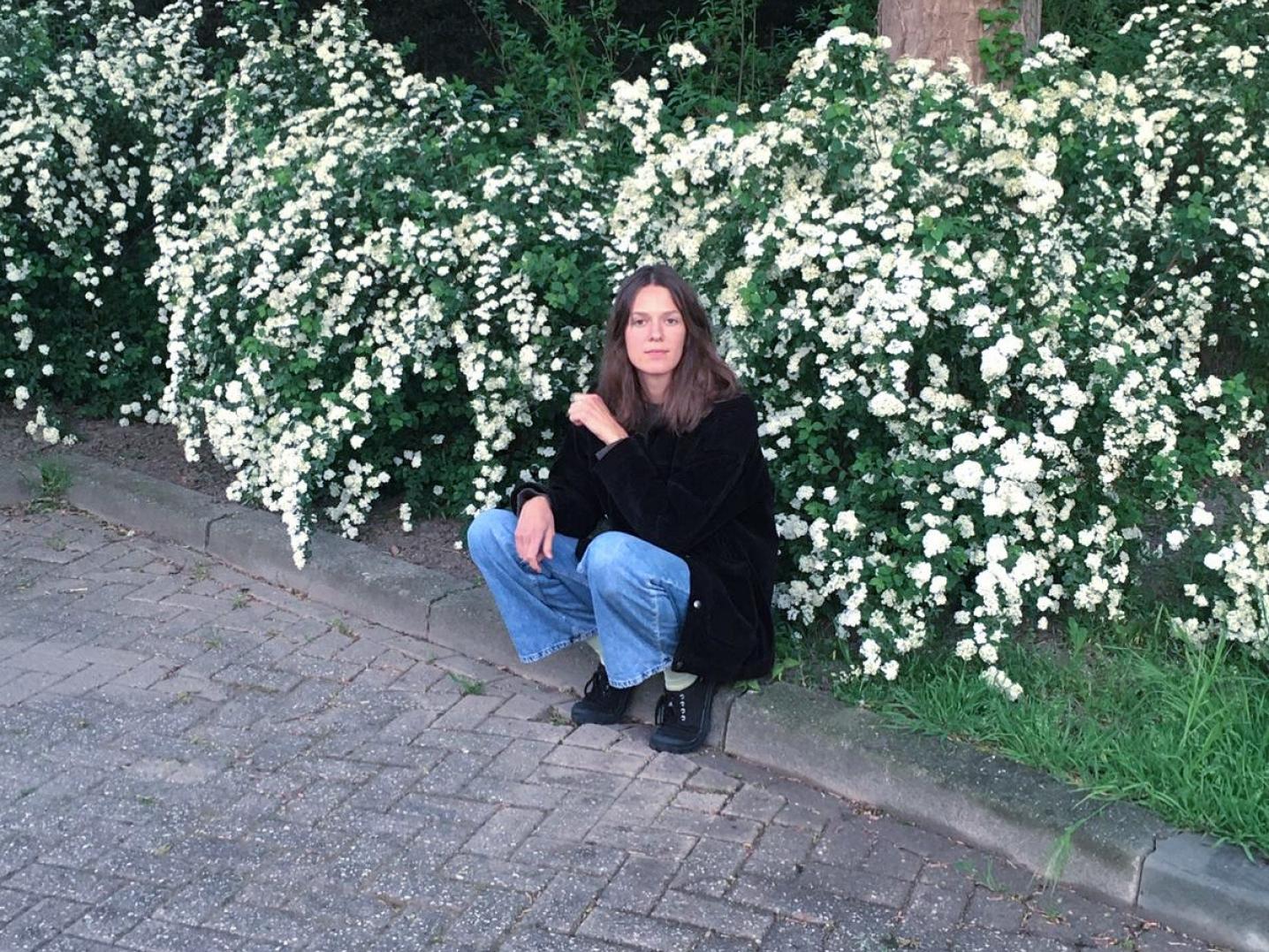 Artist Sanne Vaassen crouching in front of a bush with white flowers