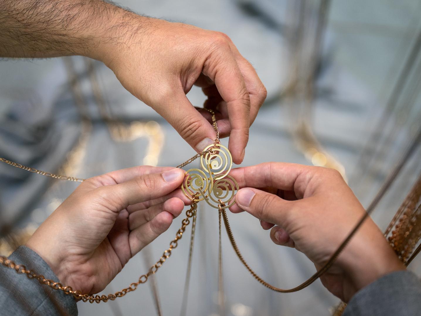 three hands holding spiral-shaped golden jewellery hanging from long golden chains