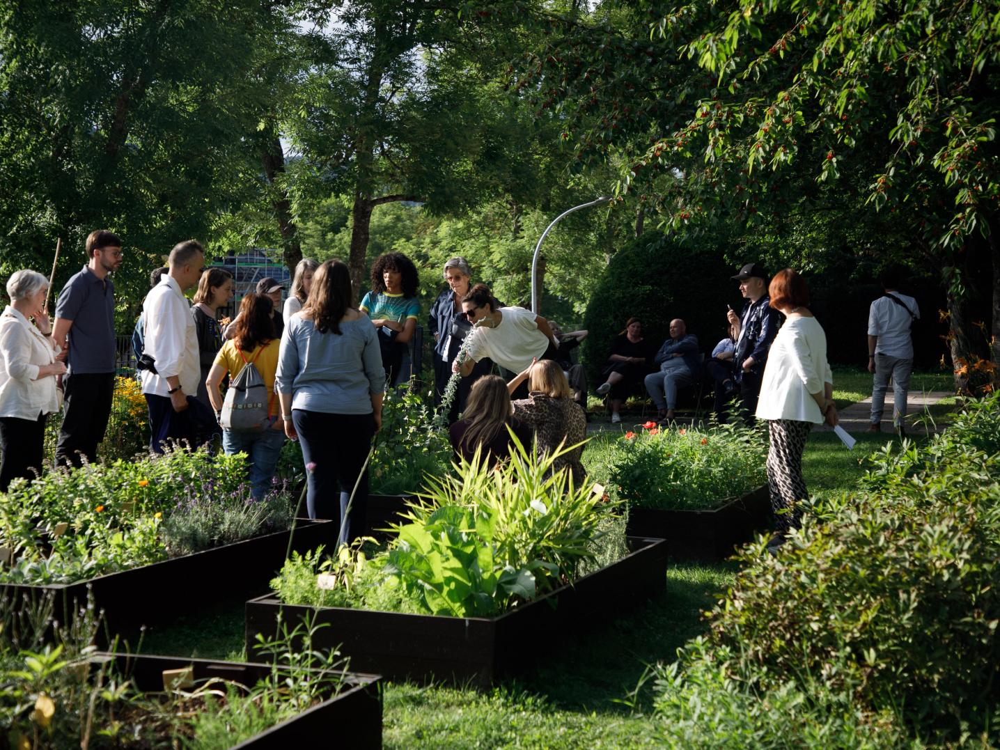 Many people standing in a public and inclusive pleasure garden including six themed herbal beds.