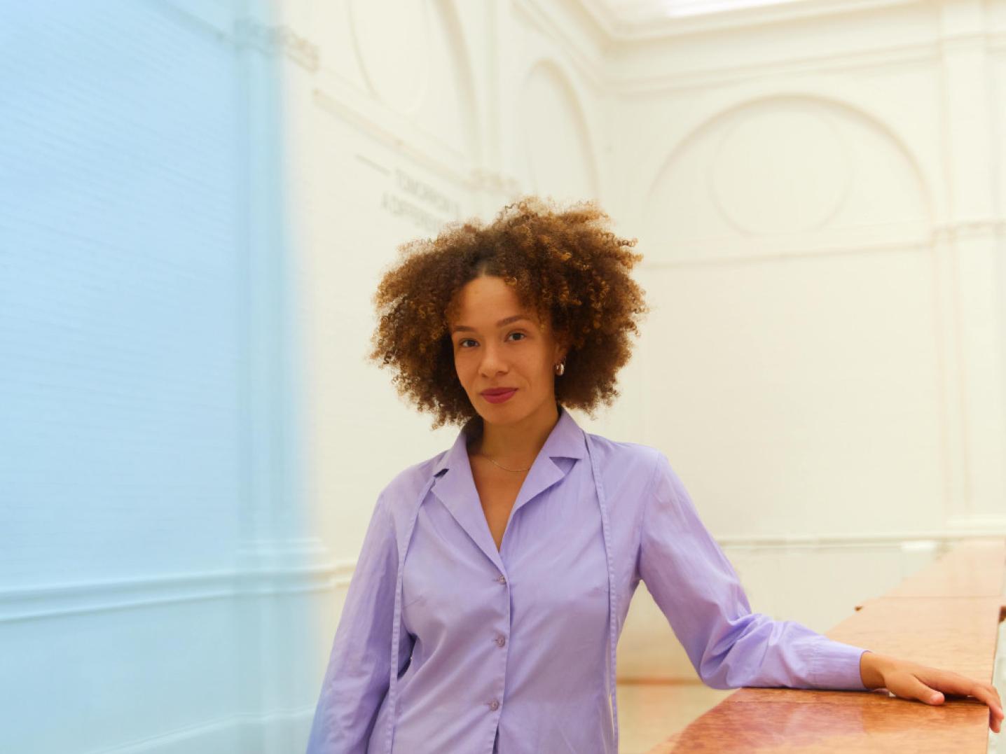Isabel Sheridan has her hand on a table standing in a well-lit white room wearing a purple blouse