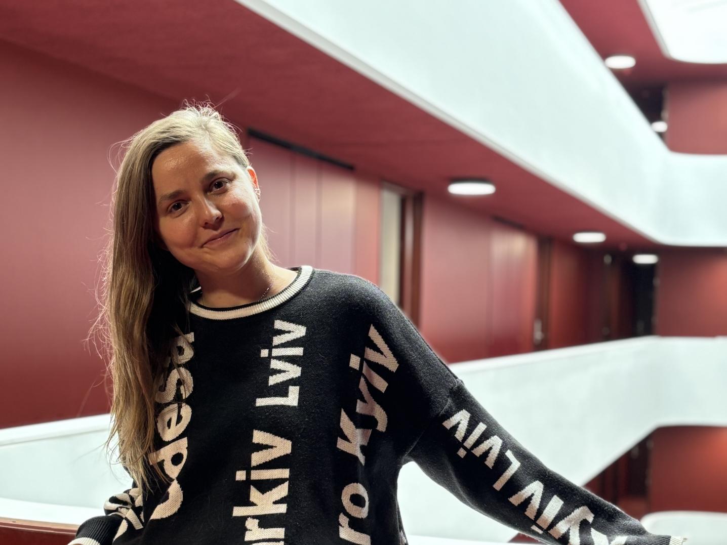 Photo of Oksana Savchuk, standing in font of burgundy soft panelling wearing a sweater that features the names of major Ukrainian cities, vertically arranged. She wears beige shorts too. 