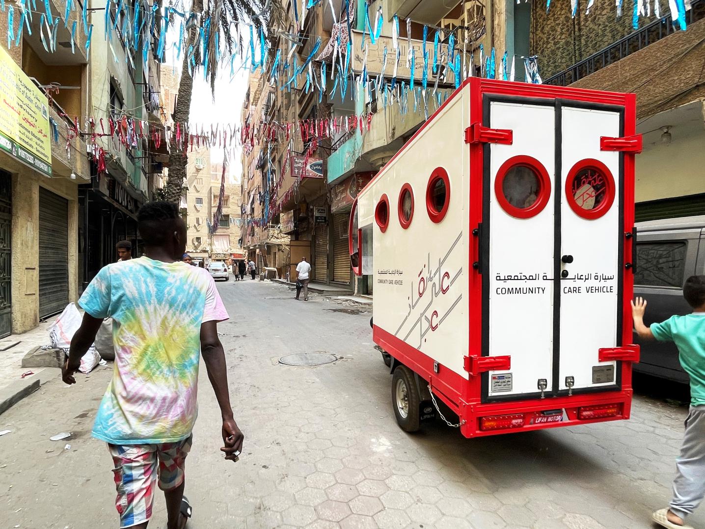 A community care vehicle, an ambulance that has been adapted to the narrow streets of an informal neighborhood of Cairo. The vehicle is parked in a street where we see several people walking both in the forefront as well as in the background. The street is decorated with garlands.