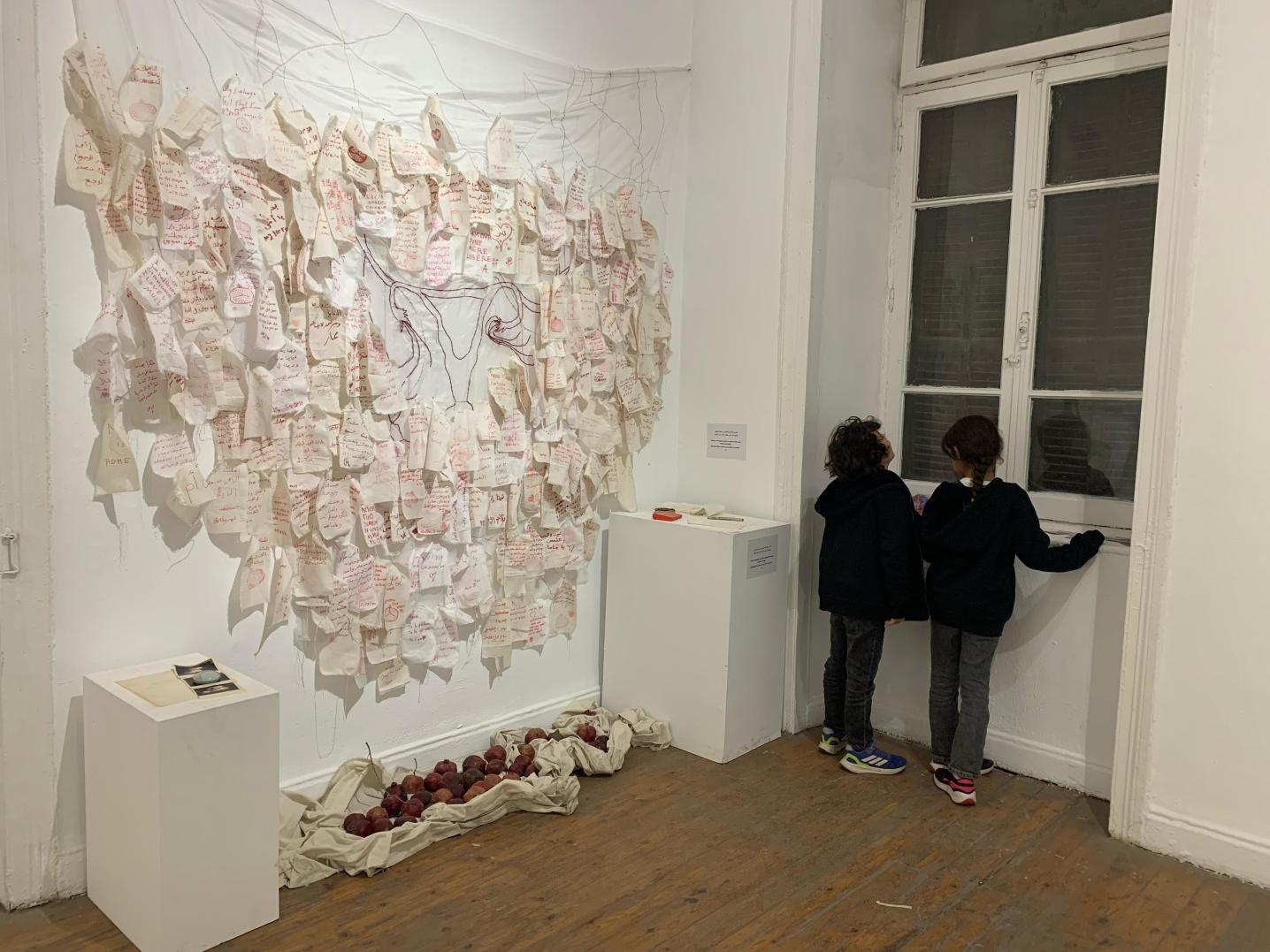 Various white sheets of fabric containing messages in red ink pinned on a large white sheet on which the image of a uterus is embroidered.