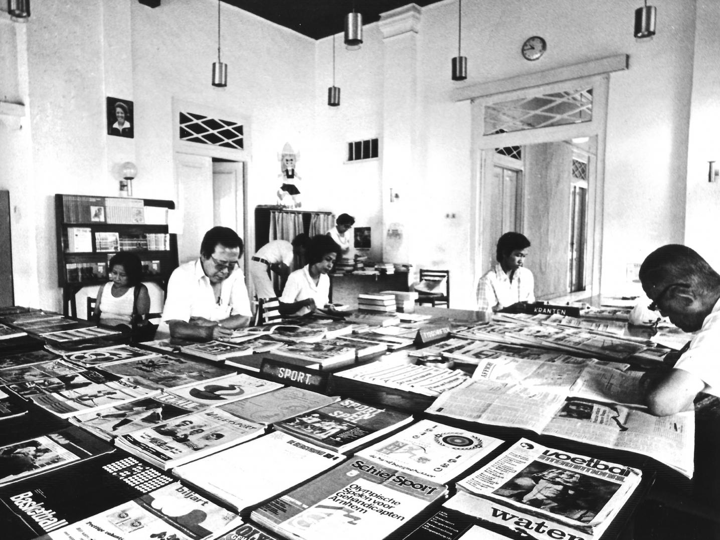 a 1970 photo of the reading room of the former Erasmus Huis, Jakarta, with people sitting around a table reading newspapers
