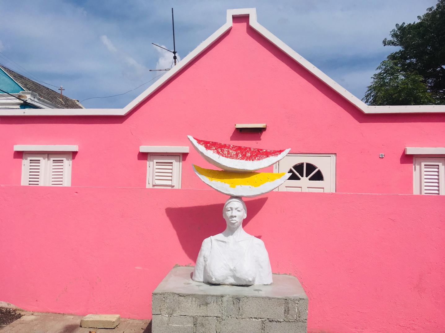 A pink building with a statue sitting in front of a woman carrying two peaces of melon on her head.