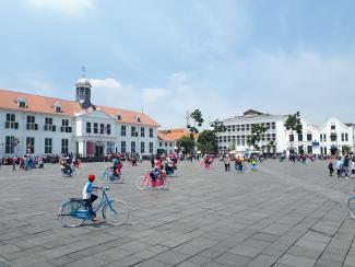 A vast square with many people, some of them on brightly coloured bikes, and buildings in the background