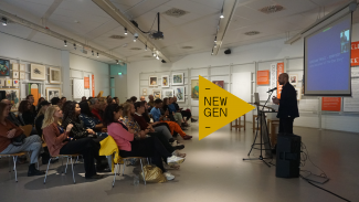 a group of seated people is listening to a man standing in front of them, there's a projection screen in the back with an image, and works of art are hanging on the wall
