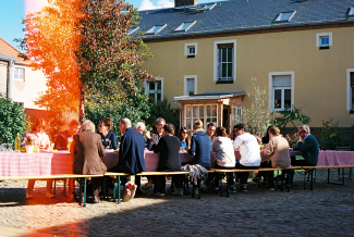 A group of people sitting at a large table outside having a lunch, in front of a yellow building