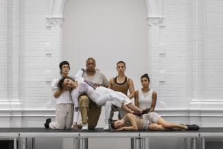 A group of people sits in front of a large white wall in the Stedelijk Museum Amsterdam. Four people are seated and two people are laying down as part of a performance.