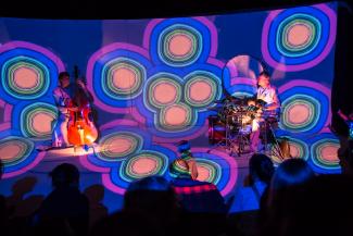 a colourful stage with a cello player and a drummer and a young audience in the foreground