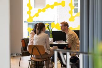 Three people in conversation sitting around a table on brown chairs. natural lighting comes in through a large window that is decorated with yellow dots