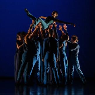 A group of dancers holding up one dancer into the sky. The single dancer's legs are raised up above, while the group supports.