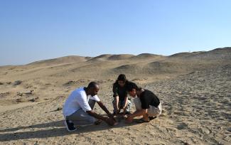 Three people are kneeling around objects on the ground that are not visible. The setting is the dessert of Saqqara, Egypt.