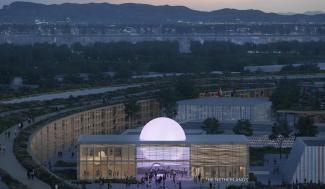 Large glass building with a light purple round shaped light on top, surrounded by other buildings in a mountain landscape