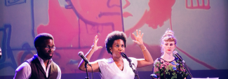 Three people on a stage with microphones and pink and purple lighting; the woman in the middle raises her hands.