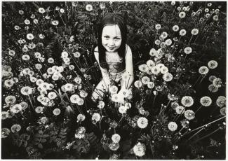 Black-and-white photo of a smiling young girl crouching in a dense field of dandelions. 