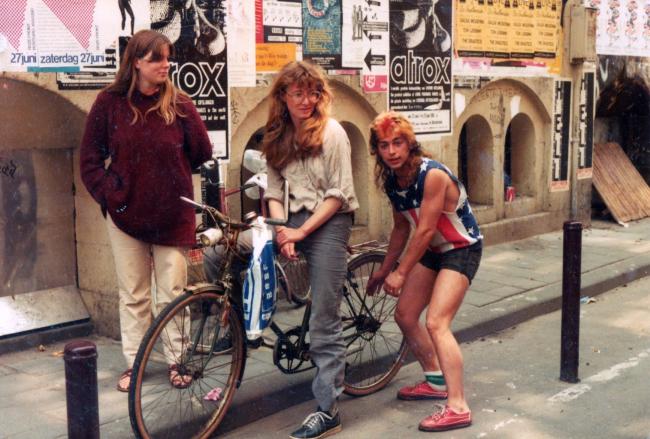 Three young people standing on a street with a bike, in front of a wall covered in posters