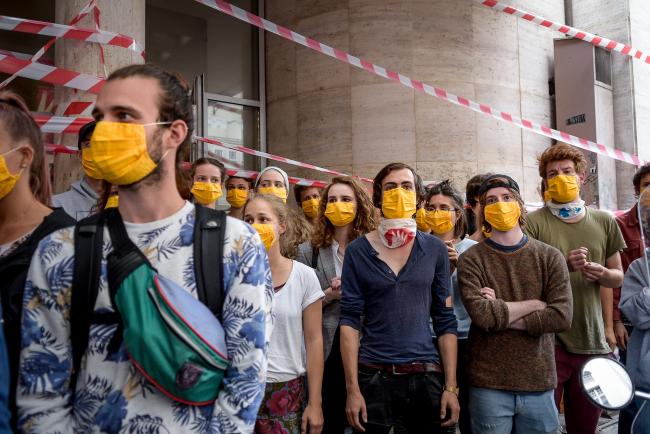 a group of young people standing in front of a building closed by plastic red-white striped ribbons, they are all wearing yellow mouth masks