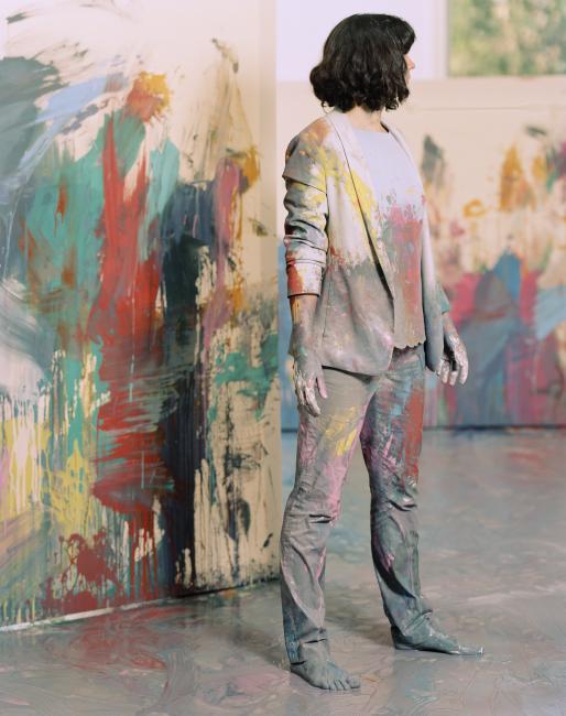 Person with short black hair standing in front of a wall smeared in green, red, yellow and black paint