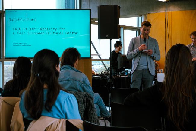 A man with a microphone next to a screen speaking in front of an audience