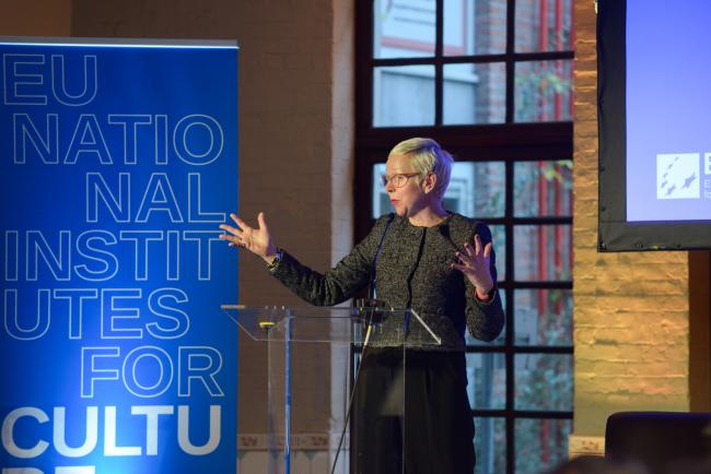 A gesturing woman giving a speech on a podium in front of a blue screen