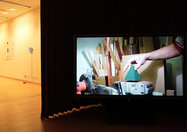 a video playing in an exhibition space, showing a hand holding a green object that is about to be placed on an installation