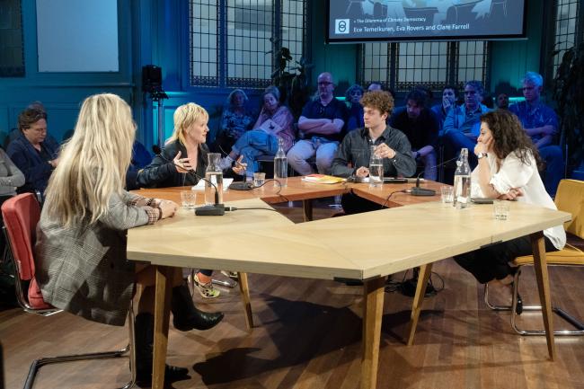 four people sitting around a table talking surrounded by an audience