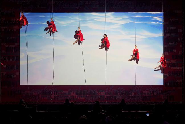 a group of 6 women in red dresses hanging from ropes on a stage with a cloudy background