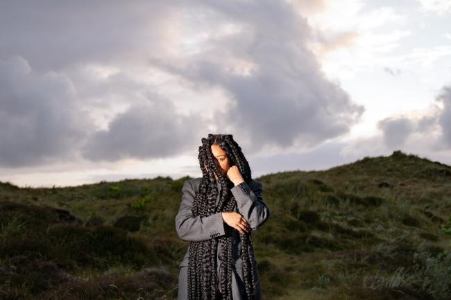 Woman with long braided black hair stands in front of green overgrown dunes on a cloudy day