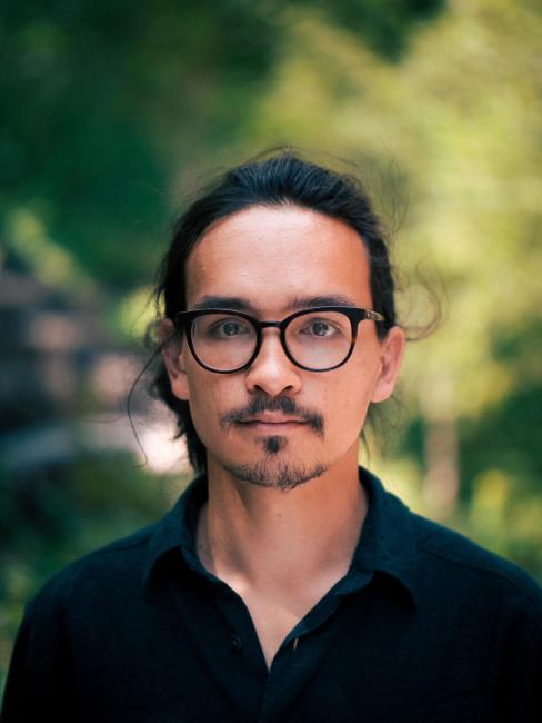 man with long black hair and black glasses calmly looks into camera while standing in green woodland