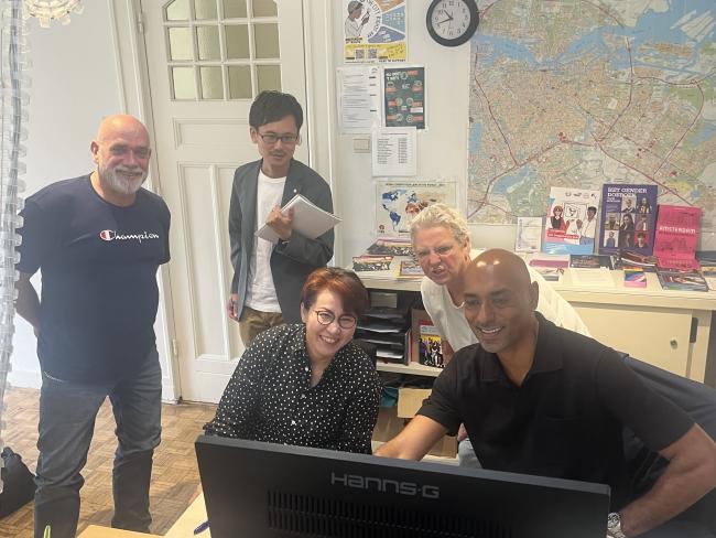five people gathered around a computer talking and smiling, a big map of the city of amsterdam in the background