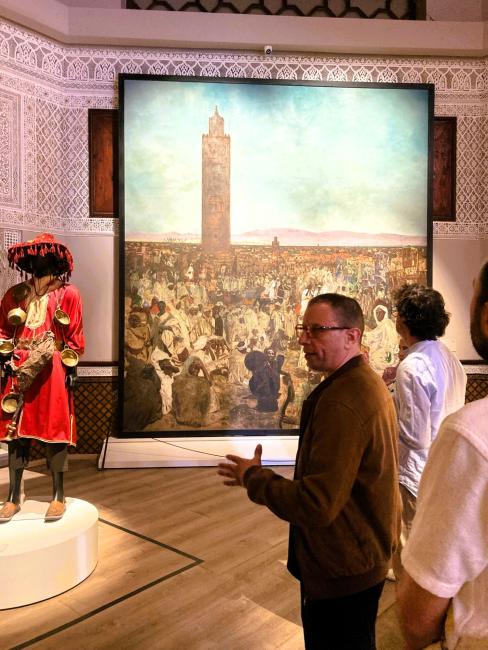 a man is standing in a museum setting talking, with a large image of a big crowded square with a tower, and a traditional moroccan costume in the background