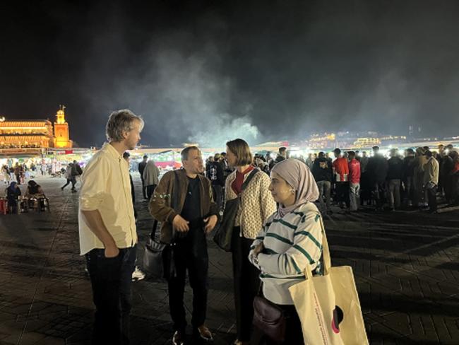Four standing on a square at night, with lit buildings and smoke in the background