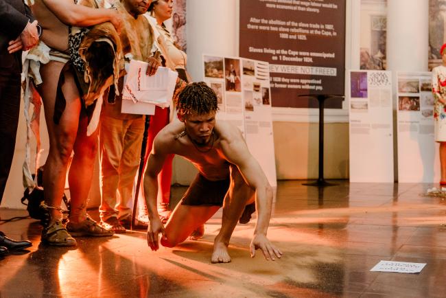 A dancer crouched on the floor, engaging in a performance. The public is gathered around, close to the performer.  surroundings are made up of informative museum pannels and the public is 