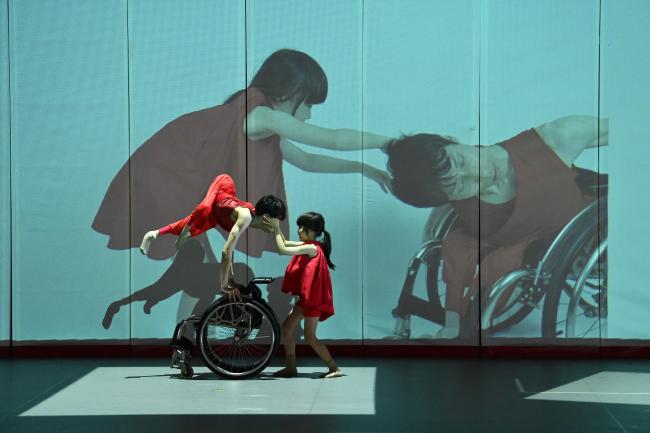 two dancers dressed in red with a wheelchair and a video of the same dancers projected on the background