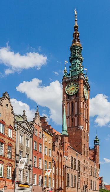 a red brick pointy tower with a bronze clock against a blue sky, and brick 17th centrury amsterdam style houses