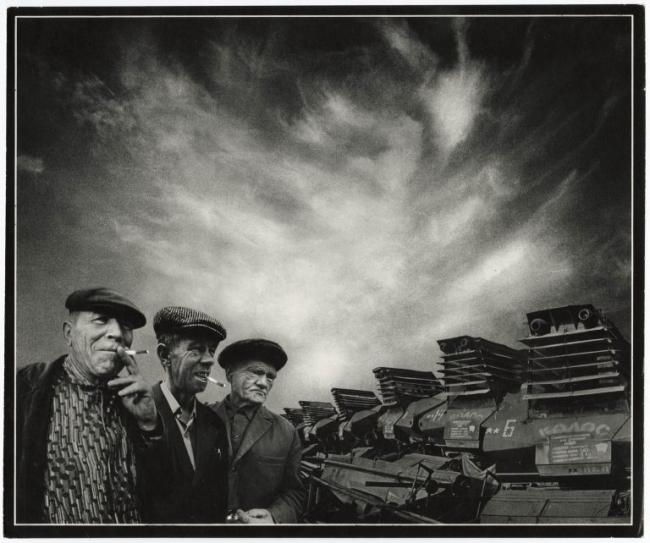 Black-and-white photo of three older men in caps standing beside large agricultural machines, under a dramatic cloud-filled sky. 