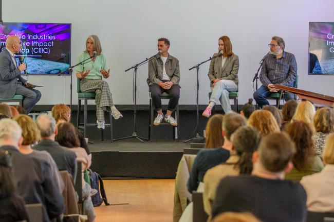 Four people sitting on a stage behind a microphone looking at the moderator on their right