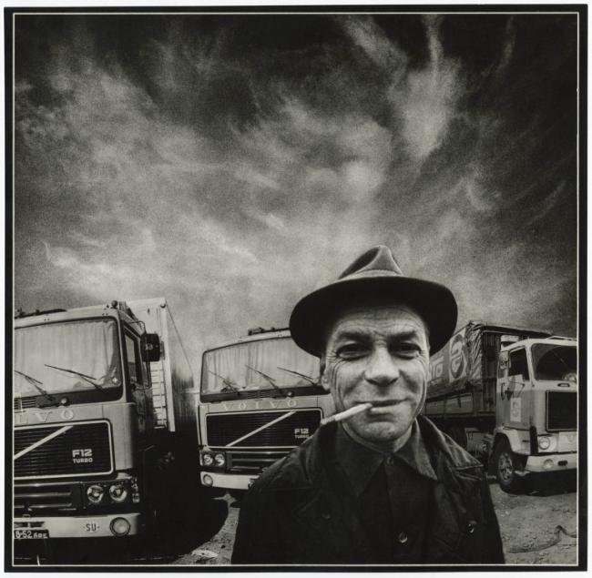 A black-and-white portrait of a man in a brimmed hat with a cigarette in his mouth, standing before three trucks under a dramatic, cloud-filled sky. 