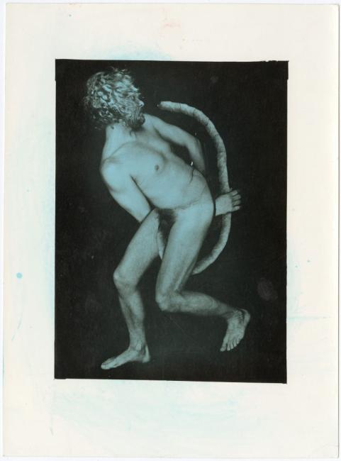 Black-and-white image of a nude adult figure with culry hair, leaning back and stepping forward while holding a long curved object against a dark background. 