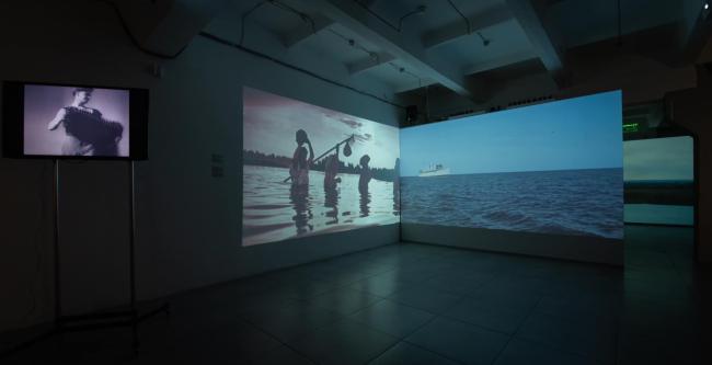 A dark gallery space with large video projections on two freestanding partition walls showing people standing in water and an open sea scene. 