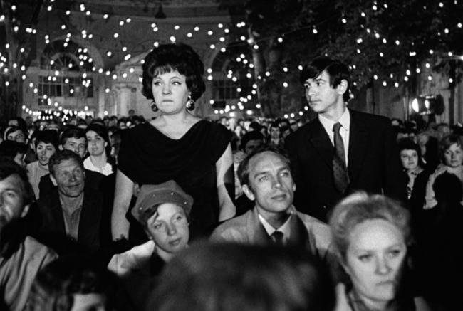 A still from the film The Long Farewell. A black and white scene of a crowded hall with festive lights. A woman stands among seated people, facing forward. 