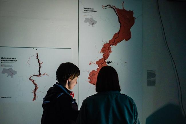 Two people seen from behind viewing a dimly lit gallery wall with the shape of the Dnipro river in red, and scientific stlyle diagrams. 